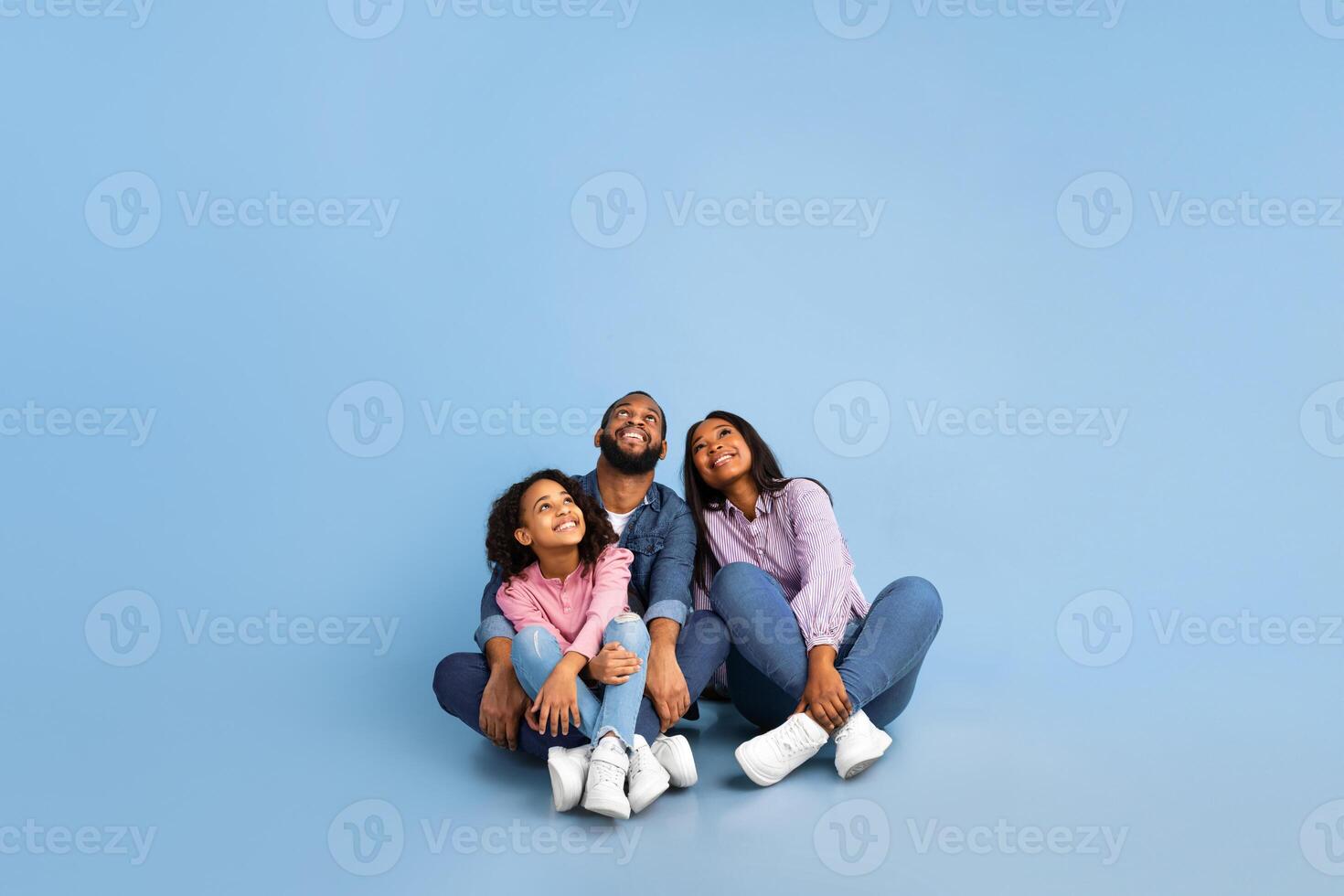 Free Place For Your Advert. Smiling African American Family Of Three People Sitting On The Floor, Looking Up At Empty Copy Space Isolated On Blue Studio Background, Vertical Shot, Banner photo