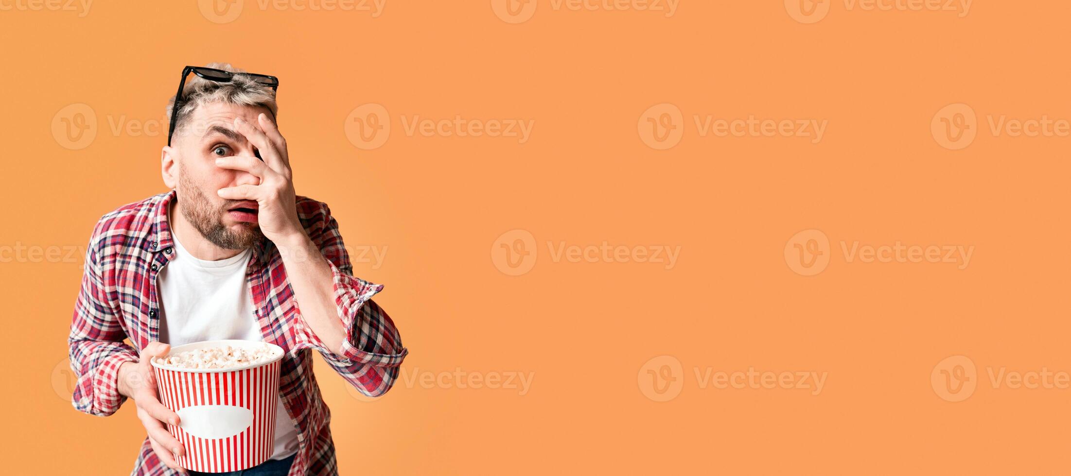 Scary movie. Shocked guy closes eyes his hand, holding bucket of popcorn, empty space photo