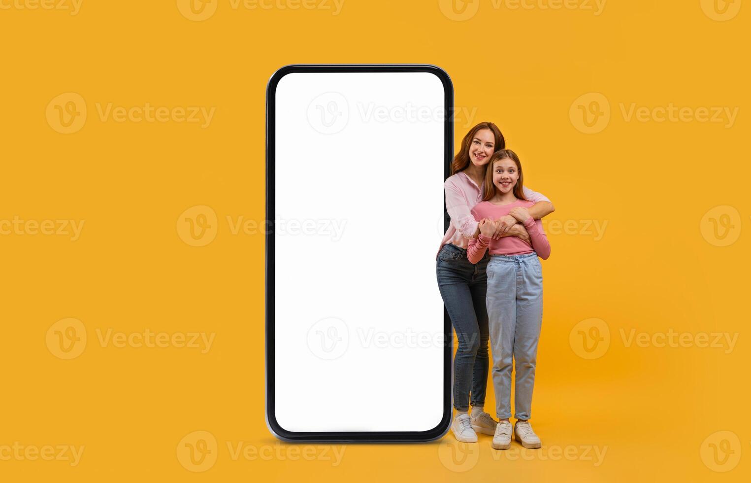 A mother and daughter are happily standing together, embracing each other, beside a large blank smartphone. The bright yellow background enhances their joyful expression. photo