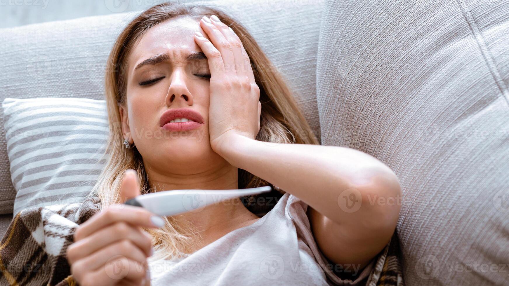 Woman has headache. She looking at thermometer and lying on sofa photo