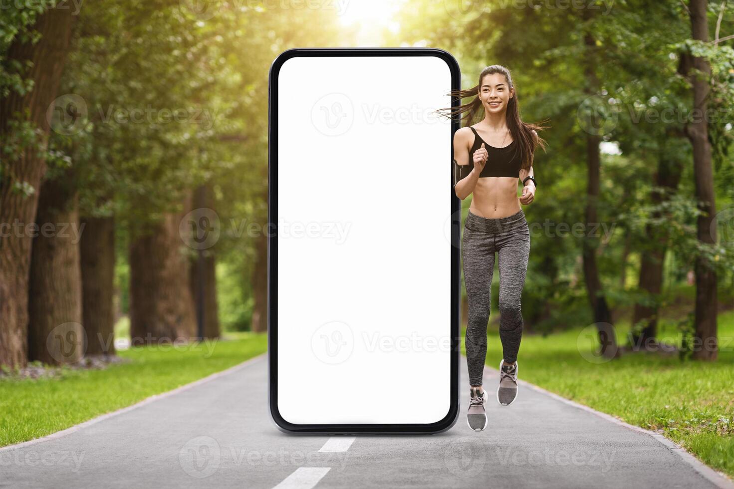 A young woman dressed in athletic gear jogs confidently along a tree-lined pathway. Beside her, a large empty smartphone display stands, framed by the bright sunlight. photo