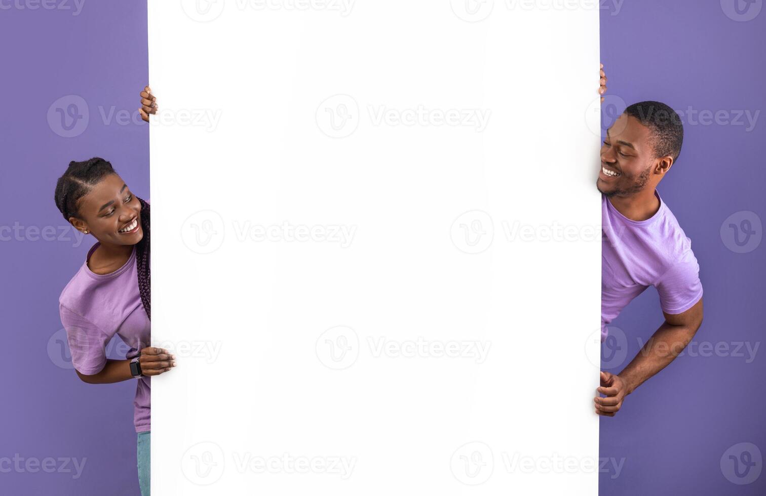 Smiling young black people holding white vertical advertisement board, demonstrating free copy space for your text or design, positive guy and lady peeking out banner, purple violet background wall photo