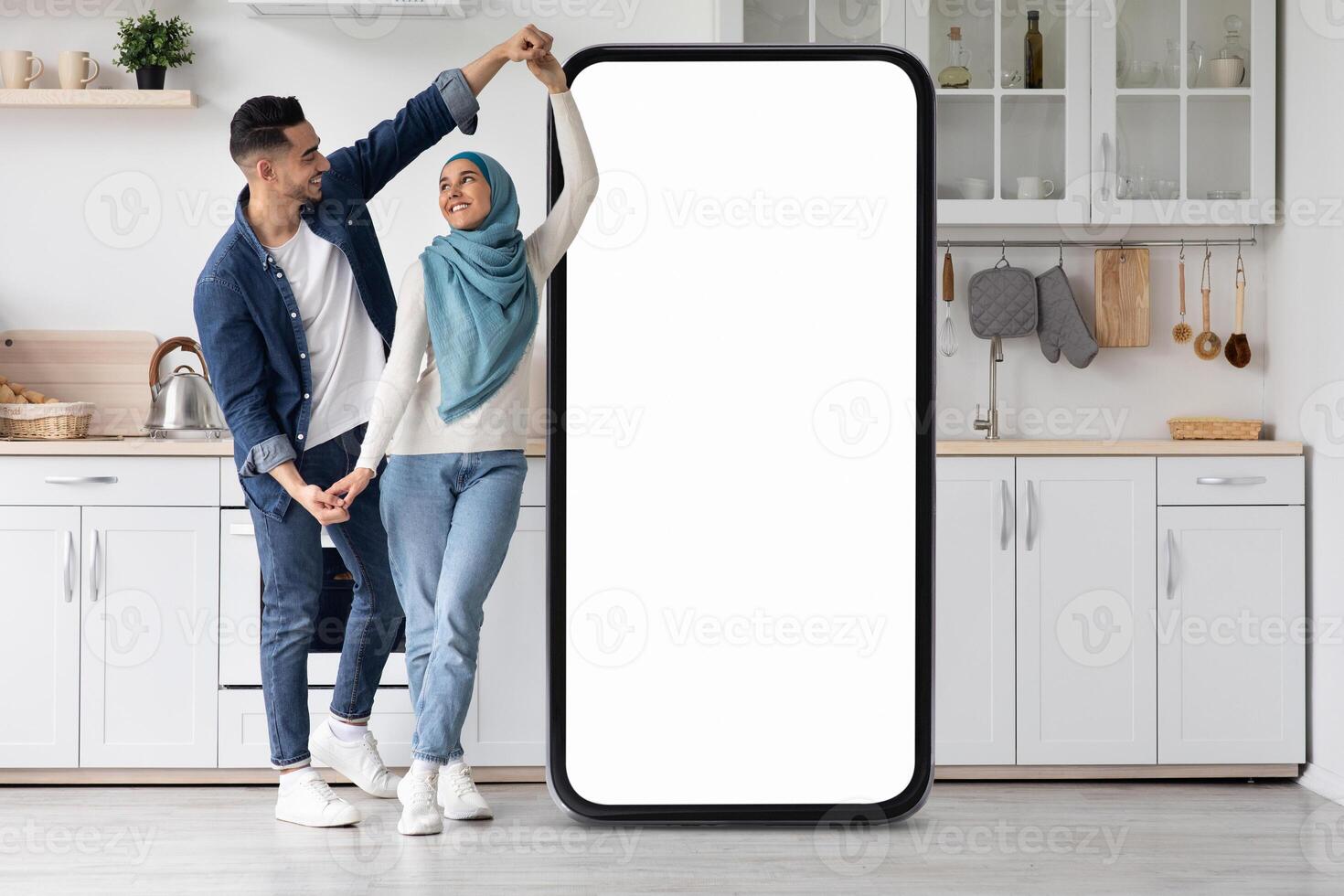 A joyful Muslim couple dances in their contemporary kitchen, playfully interacting around a large smartphone frame while enjoying their time together in a bright, inviting space. photo