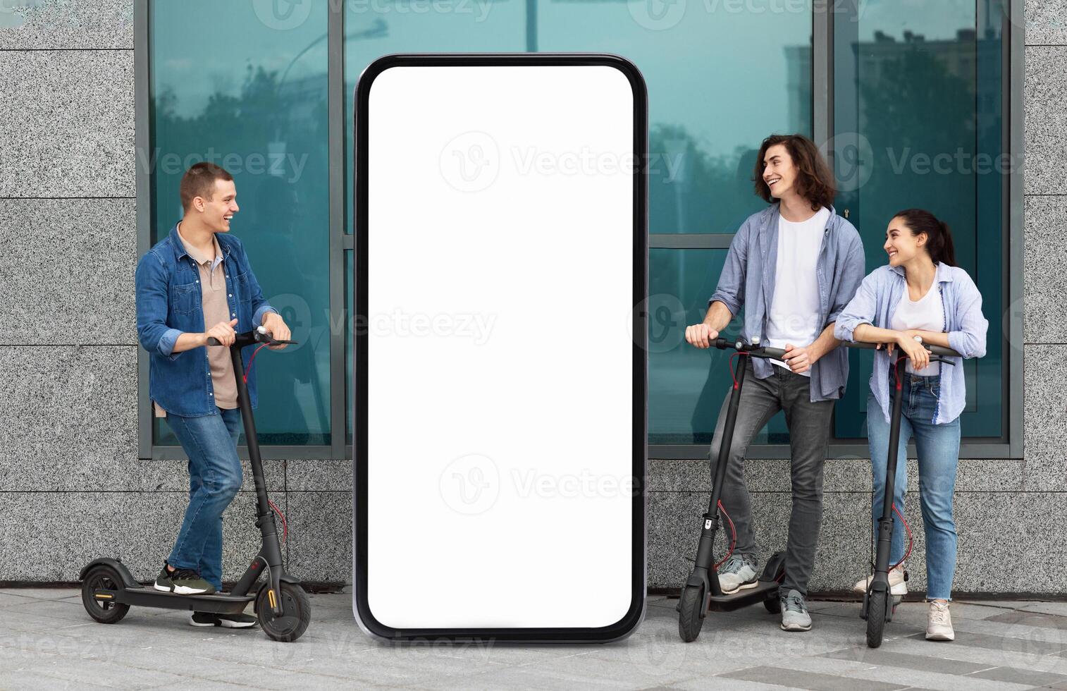 A group of three young adults stands alongside electric scooters, chatting and laughing near a large blank smartphone display on a city street during a sunny day. photo