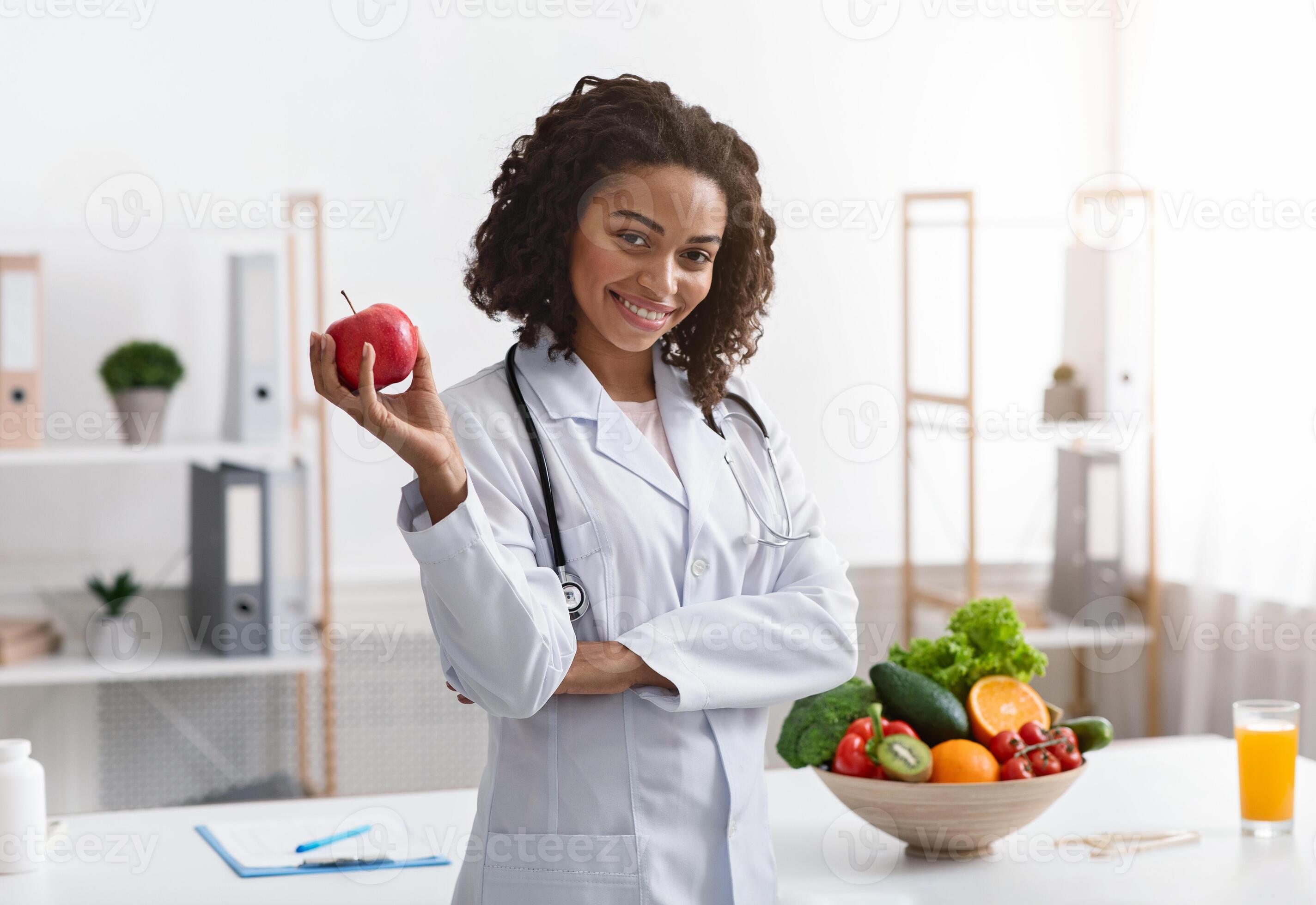 Positive black woman nutritionist posing at clinic holding apple copy Positive black woman nutritionist posing at clinic holding apple copy