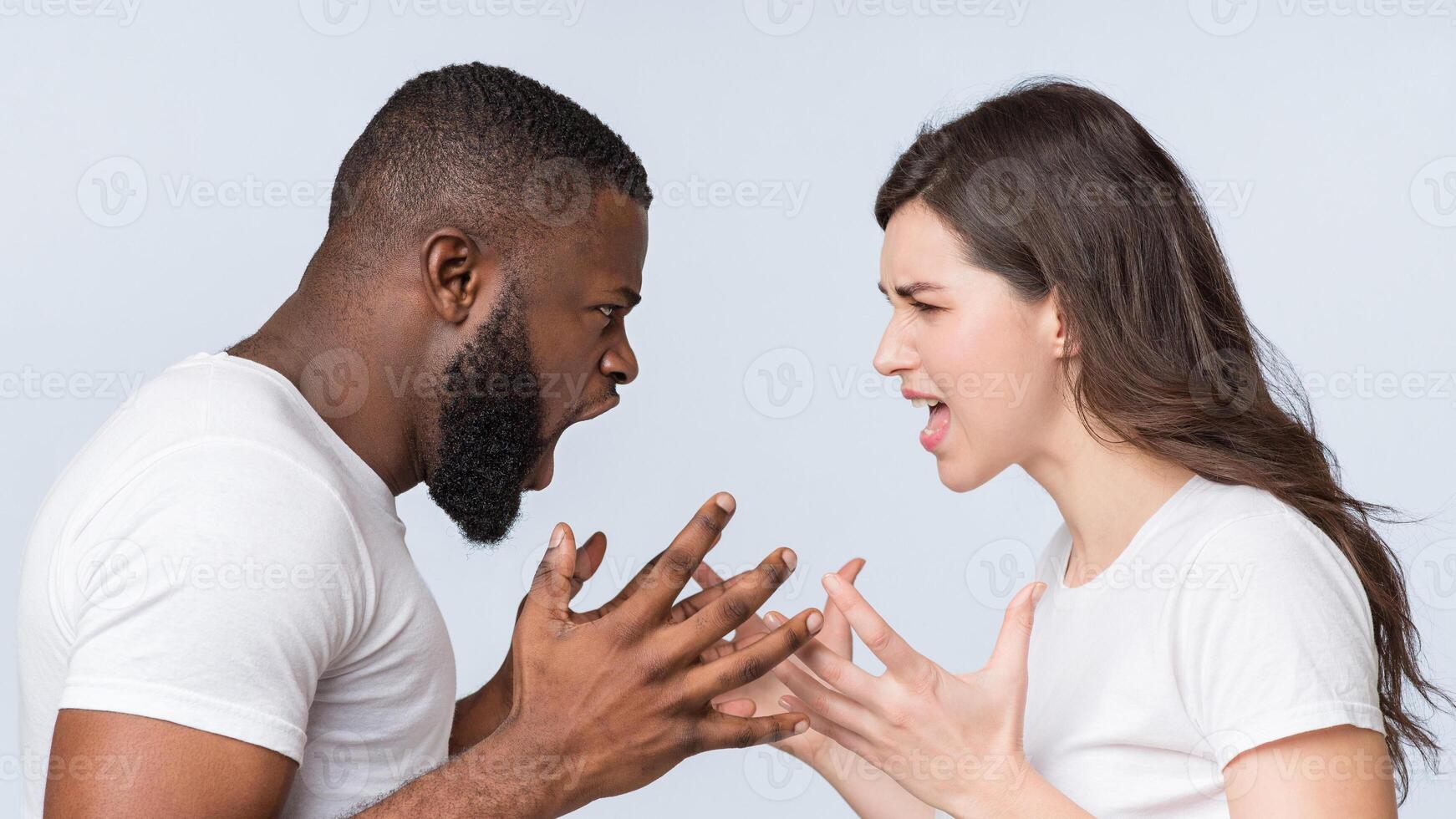 Multiracial couple quarreling, shouting at each other, having relationship problems, standing together over light background, side view photo