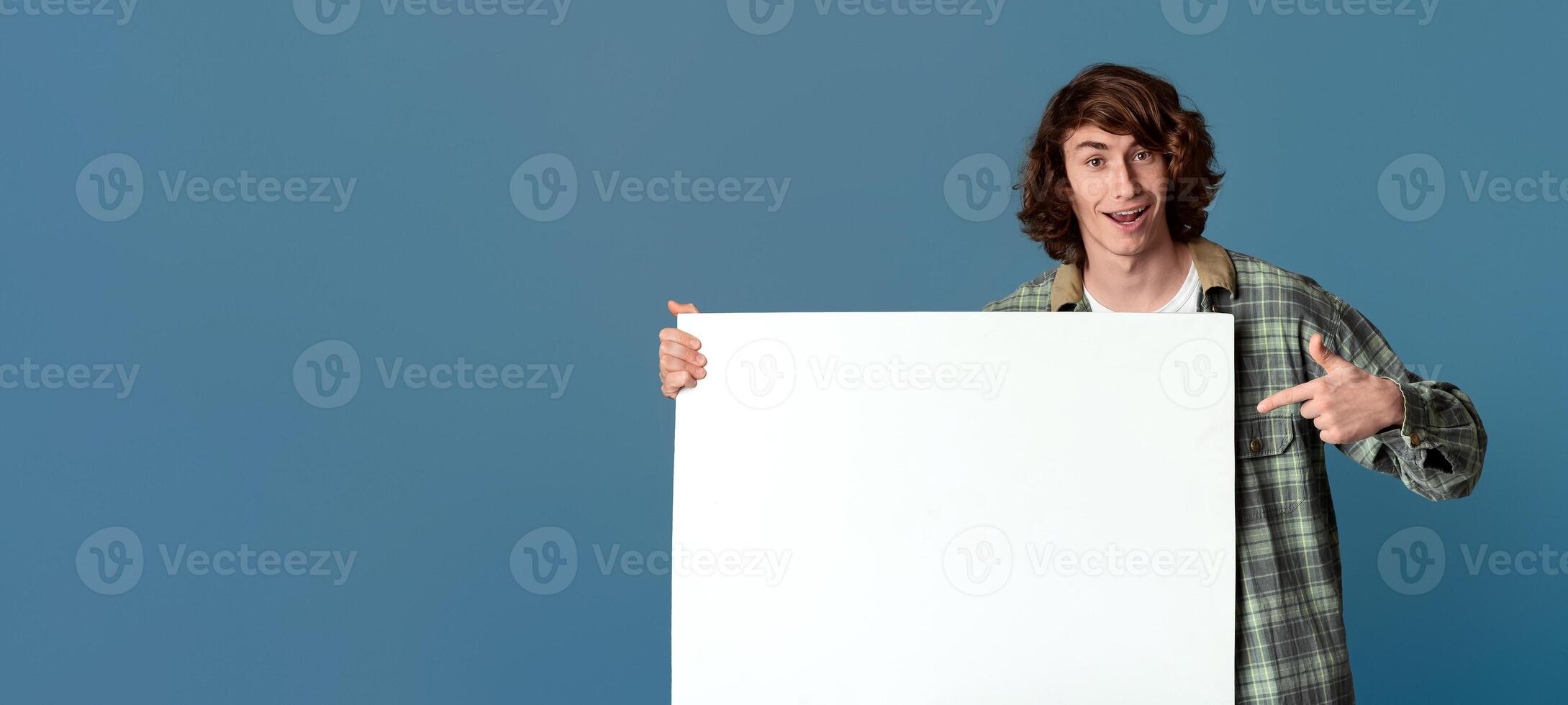 Funky millennial guy pointing at poster with empty space for your ad on turquoise background photo