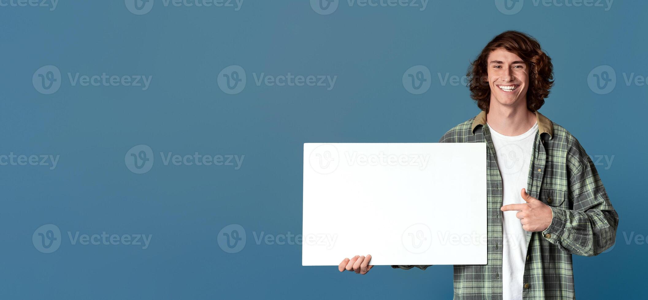 Joyful millennial guy pointing at blank poster on turquoise background, space for your design photo