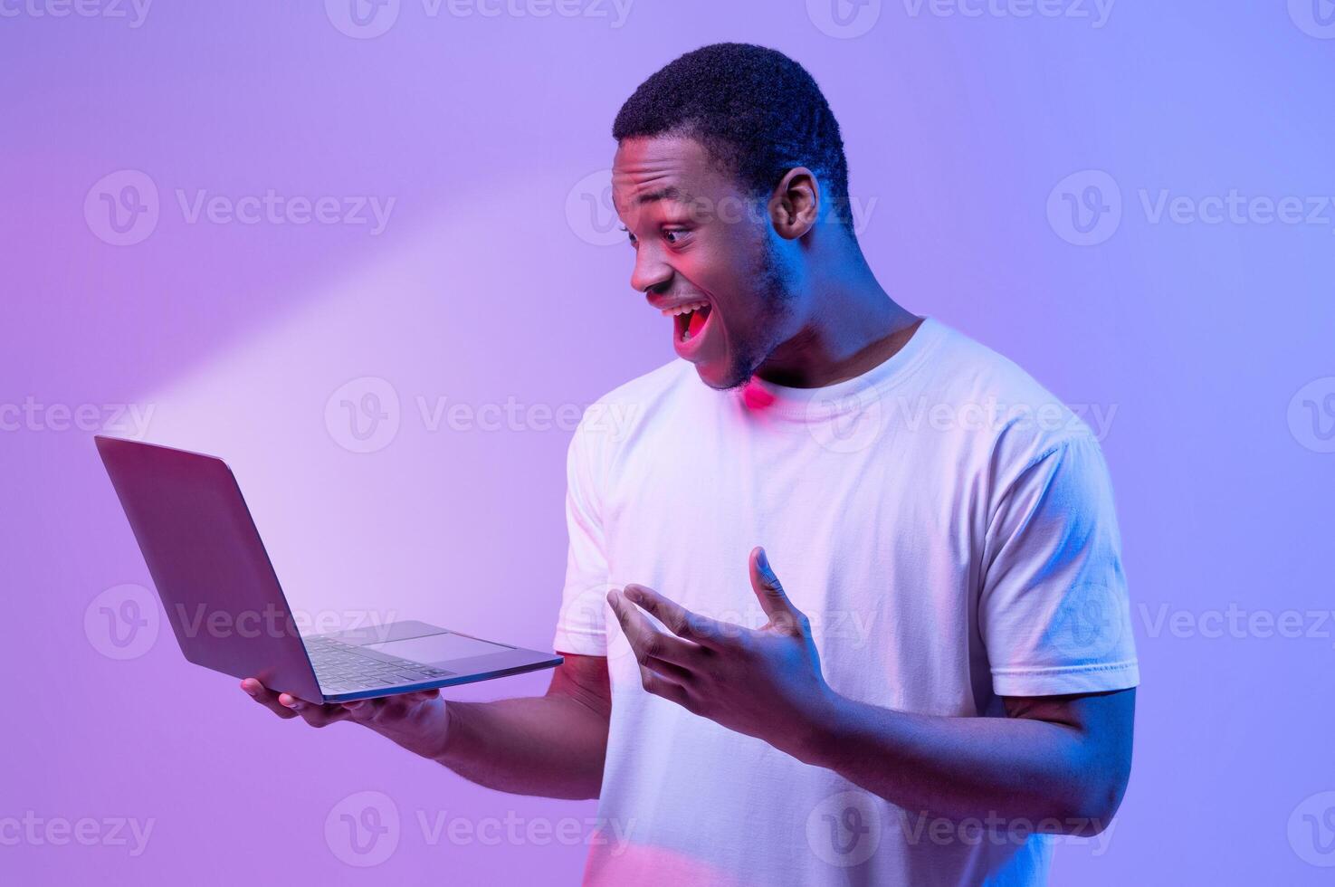 Online Offer. Surprised African American Man Holding Laptop Looking At Glowing Screen, Excited Black Guy Playing Games Or Browsing Internet On Computer, Stranding In Neon Light Over Purple Background photo