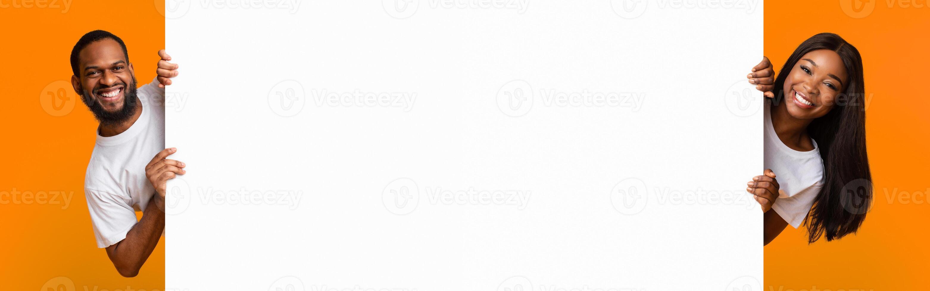 Promotion Billboard Concept. Cheerful African American Guy And Lady Holding White Advertisement Board, Standing Together On Yellow Background, Looking At Camera Peeping Out The Two Sides Of Sign Board photo