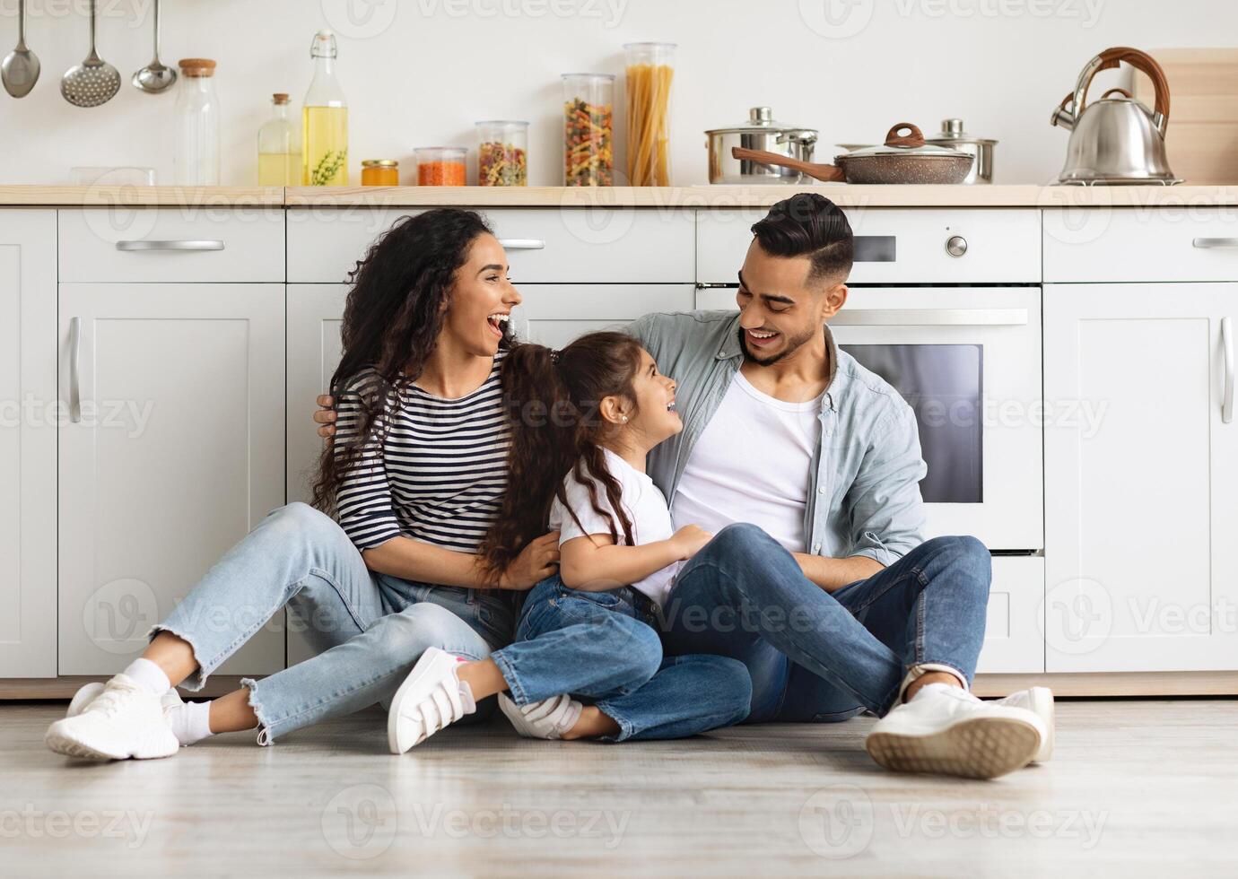 Happy arab family spending weekend together at home, cheerful middle-eastern young father, mother and cute little girl daughter sitting on tile floor at white kitchen, having conversation, copy space photo