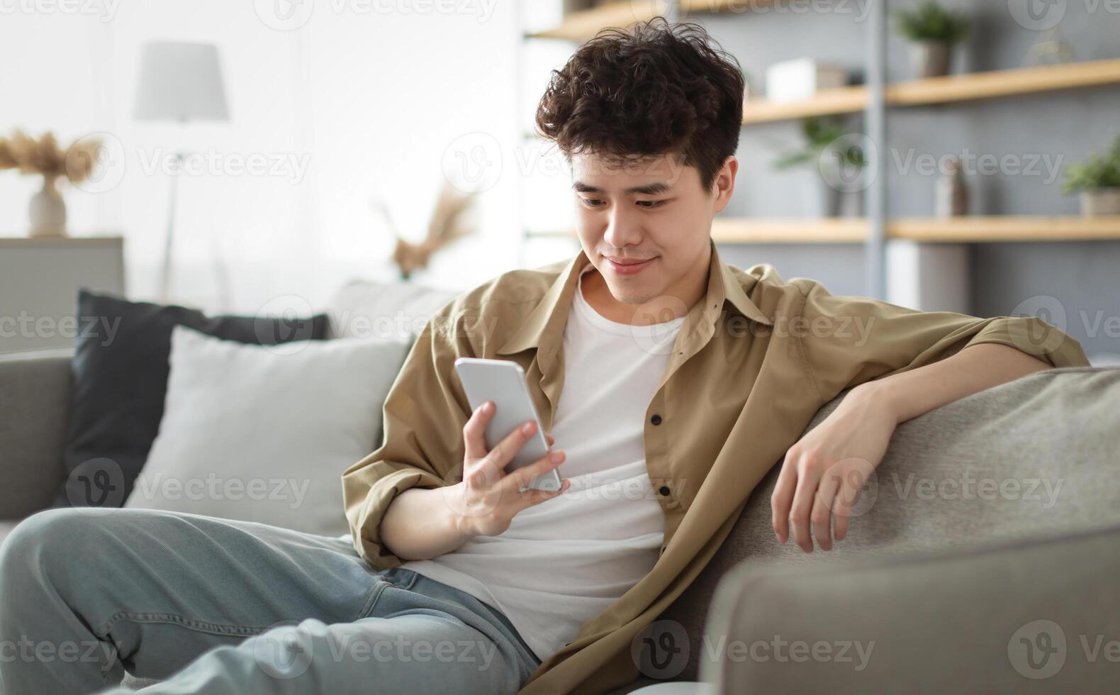 Enjoying Weekend. Handsome Relaxed Asian Man Using Smartphone While Sitting Leaning On Couch At Home, Happy Guy Resting In Living Room, Browsing Internet Or Messaging With Friends, Selective Focus photo