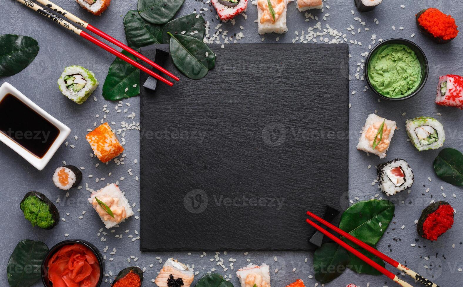 Top view on traditional japanese empty square black slate plate framed with sushi, ginger, soy, wasabi bowls, green leaves and chopsticks on rustic grey backround copy space photo