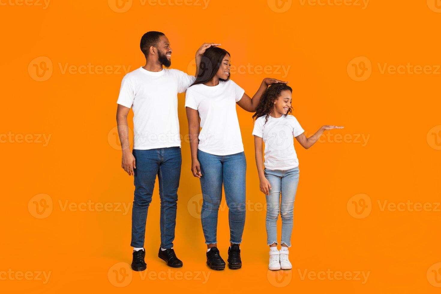 Full body length size photo of three members of family measuring height with hand palm. Smiling mum, dad and daughter cheking growing up development standing isolated on yellow orange color background