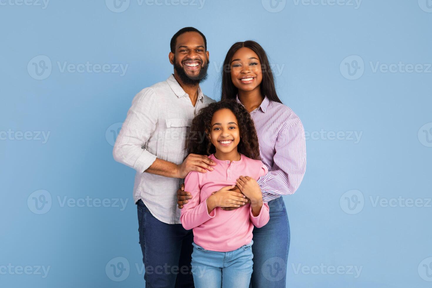 Love And Care Concept. Portrait of African American mum and dad embracing smiling daughter standing isolated on blue studio background. Cheerful man and woman posing with girl and looking at camera photo