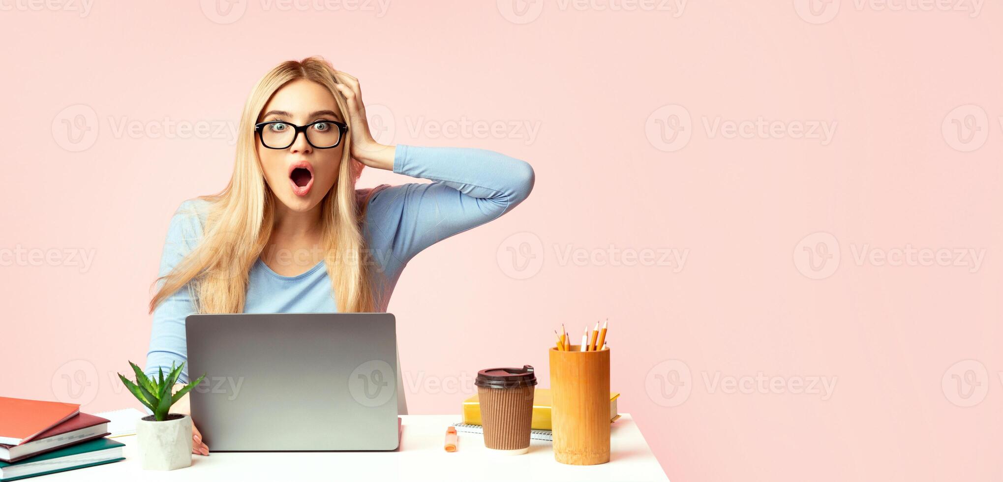 Deadline Concept. Surprised woman in glasses sitting at desk and holding hand on head, pink studio wall, copyspace photo