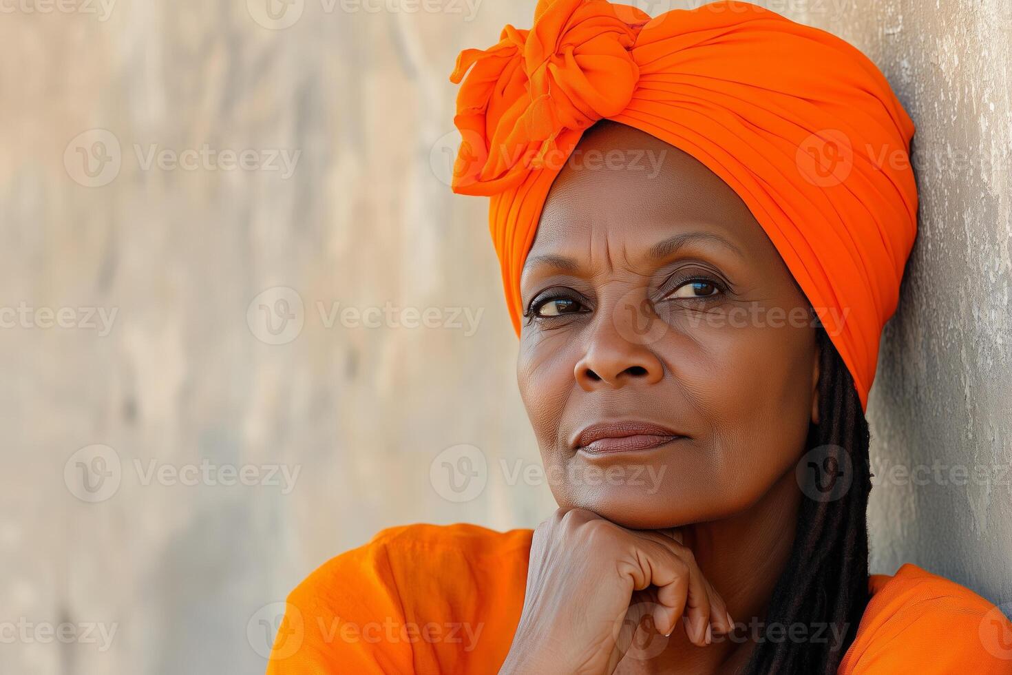 An older woman with long dreadlocks and an orange head wrap resting thoughtfully against a textured wall outdoors photo
