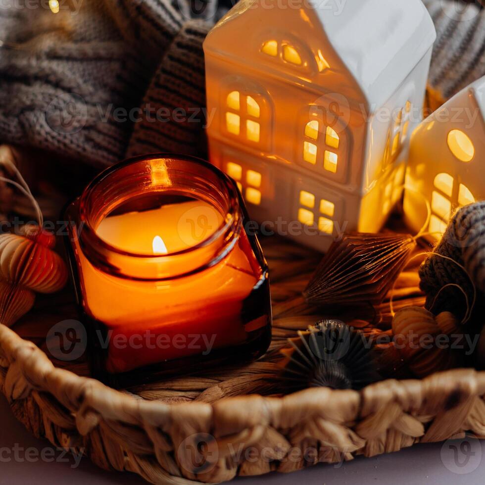 Winter or autumn cozy interior decorations. Burning candle in jar, white decorative ceramic houses, gray cozy sweater and small paper honeycomb baubles. Christmas cozy winter home decor. photo