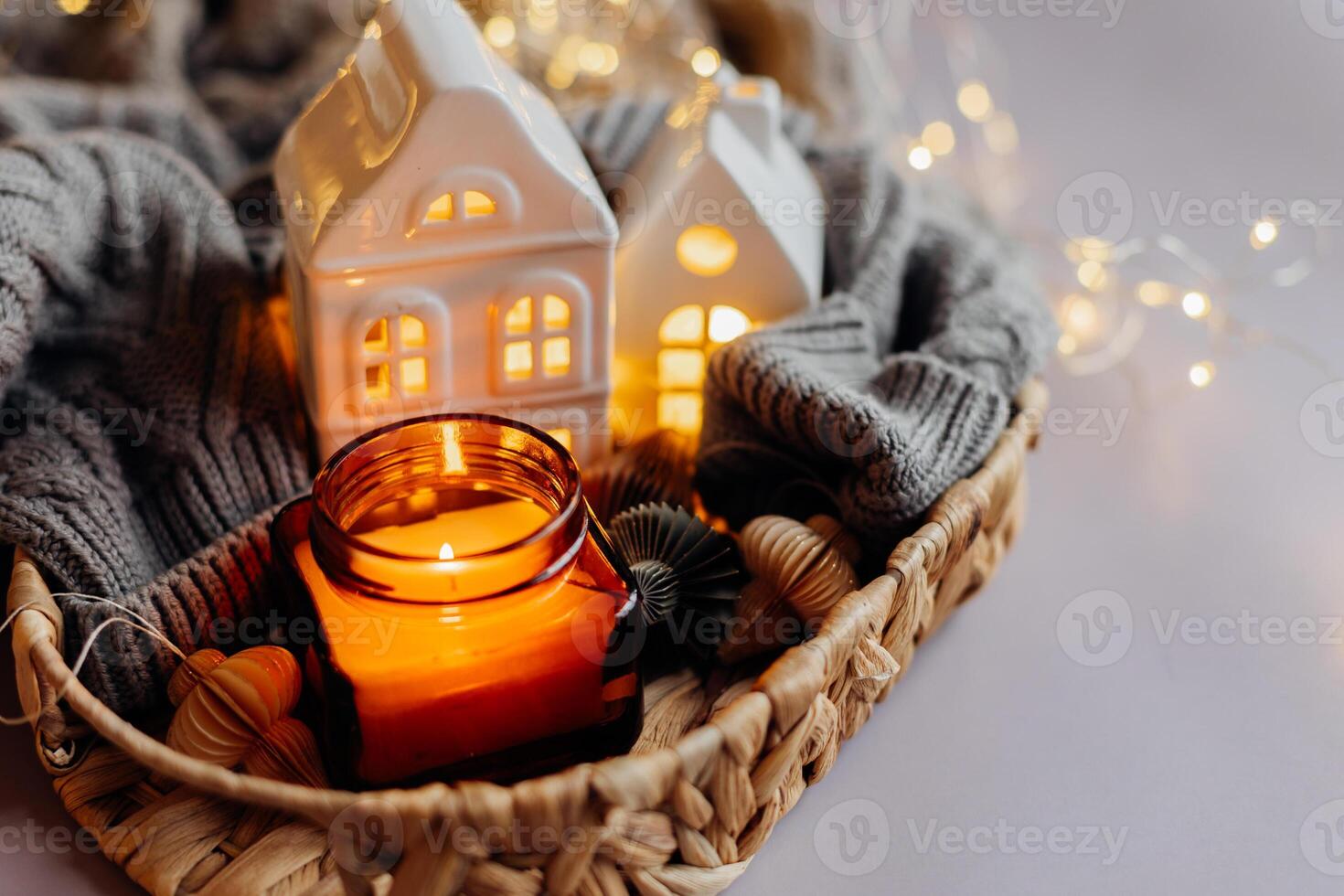 Winter or autumn cozy interior decorations. Burning candle in jar, white decorative ceramic houses, gray cozy sweater and small paper honeycomb baubles. Christmas cozy winter home decor. photo