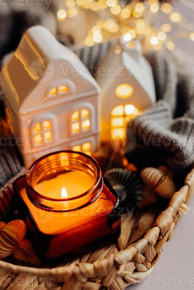 Winter or autumn cozy interior decorations. Burning candle in jar, white decorative ceramic houses, gray cozy sweater and small paper honeycomb baubles. Christmas cozy winter home decor. photo