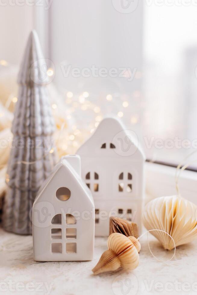 New year interior decorations. White decorative ceramic houses, Christmas tree, garland lights, paper ornate on background. Christmas cozy winter home decor. photo