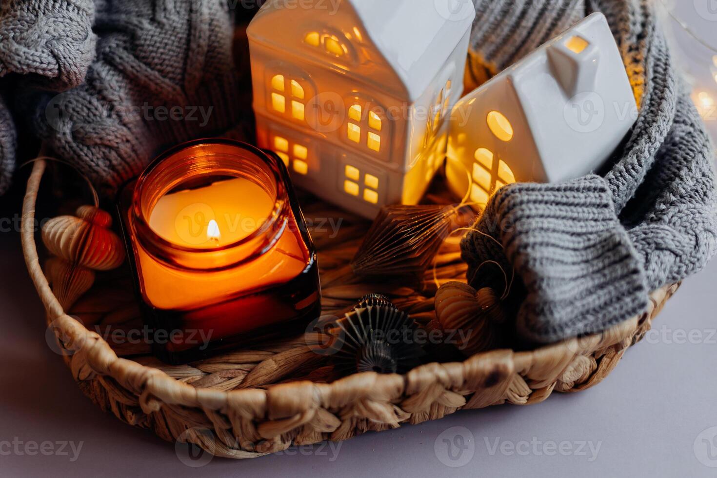 Festive stylish decoration. Burning candle in jar, white decorative ceramic houses, gray cozy sweater and small paper honeycomb baubles. photo