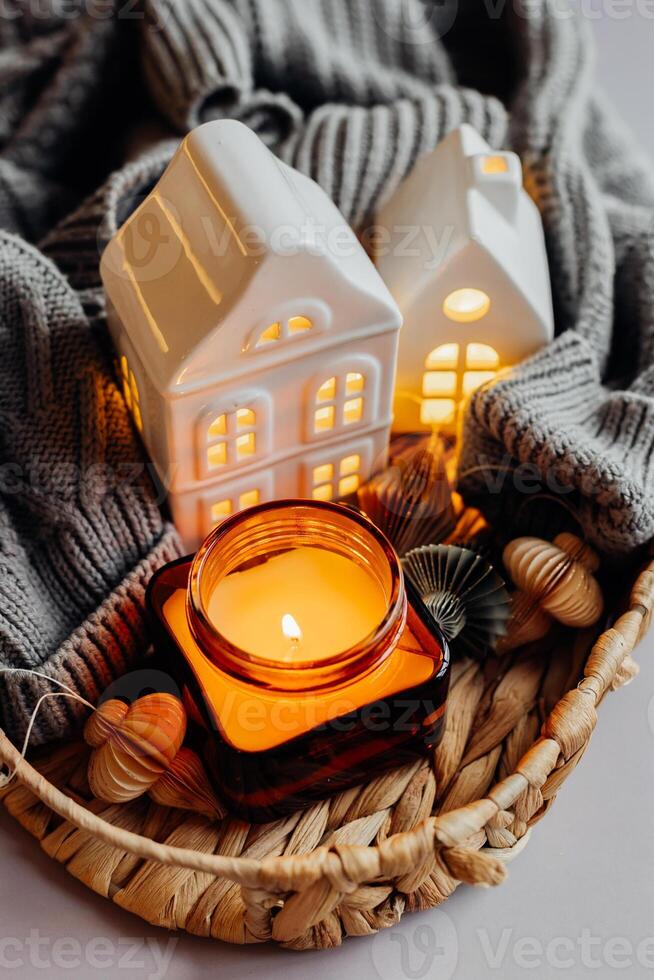 New year cozy interior decorations. Burning candle in jar, white decorative ceramic houses, gray cozy sweater and small paper honeycomb baubles. Christmas cozy winter home decor. photo