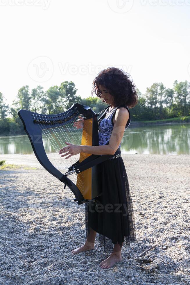 beautiful curly haired woman in fairytale clothes playing a small harp relaxing on a beach along the river at sunset photo