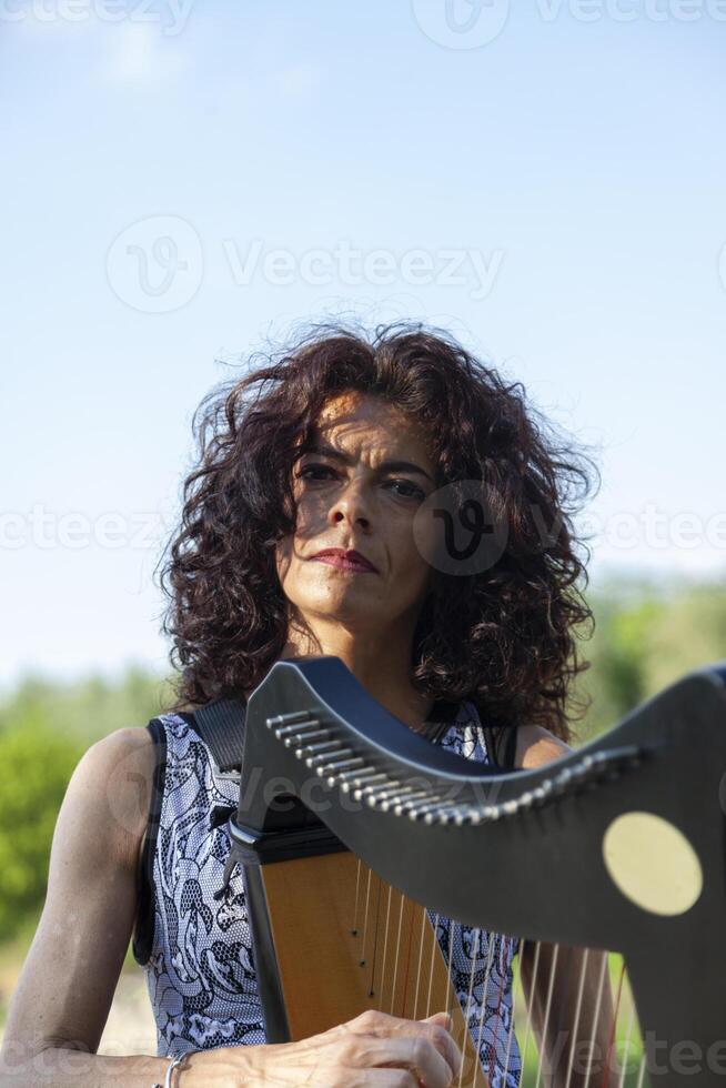 beautiful curly haired woman in fairytale clothes playing a small harp relaxing on a beach along the river at sunset photo