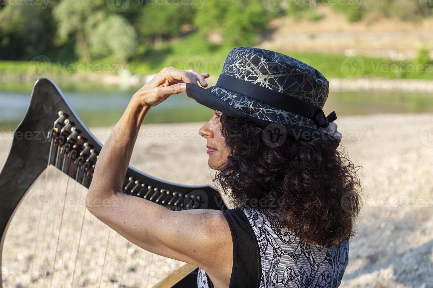 beautiful curly haired woman in fairytale clothes playing a small harp relaxing on a beach along the river at sunset photo