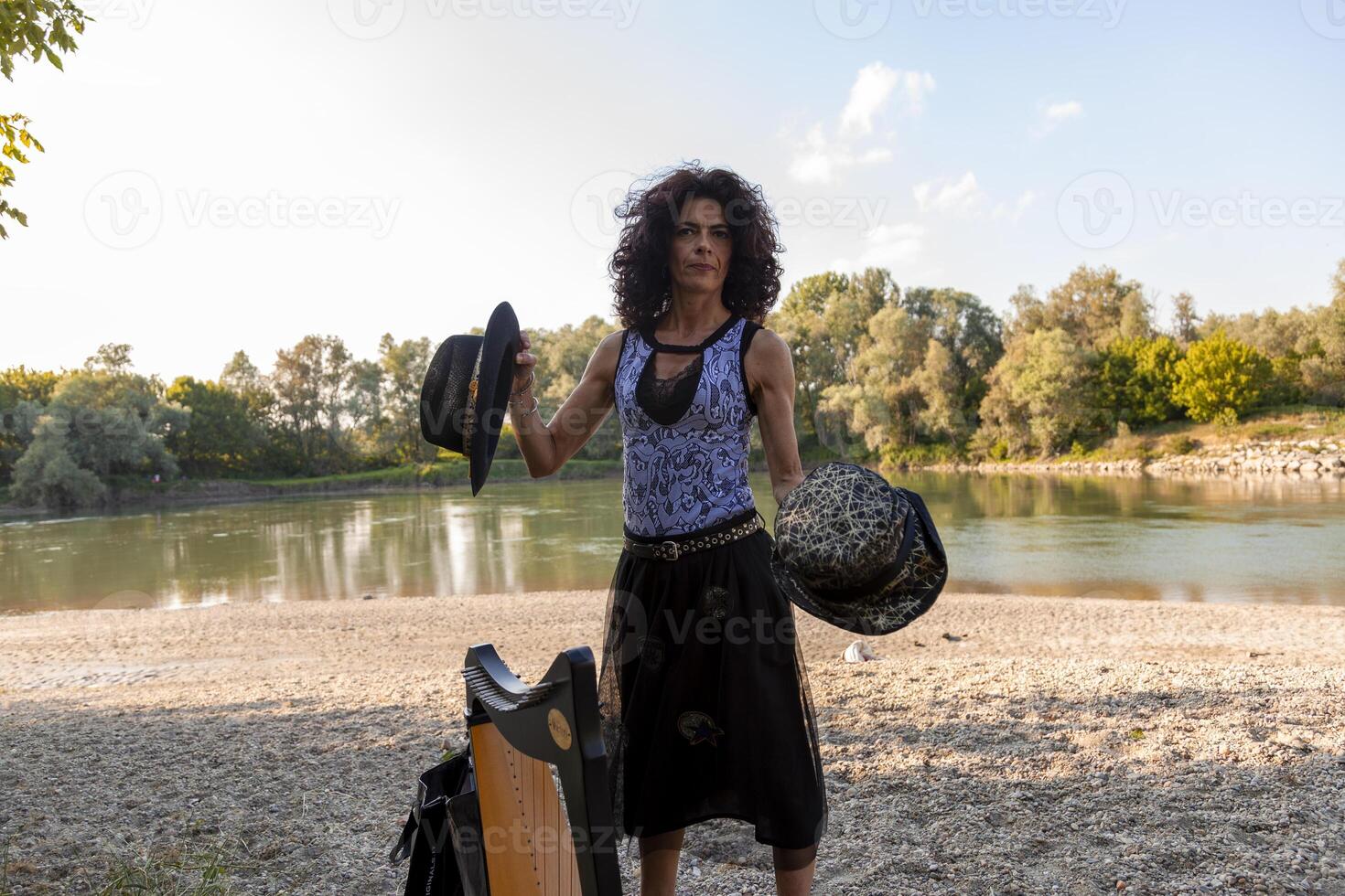 beautiful curly haired woman in fairytale clothes playing a small harp relaxing on a beach along the river at sunset photo