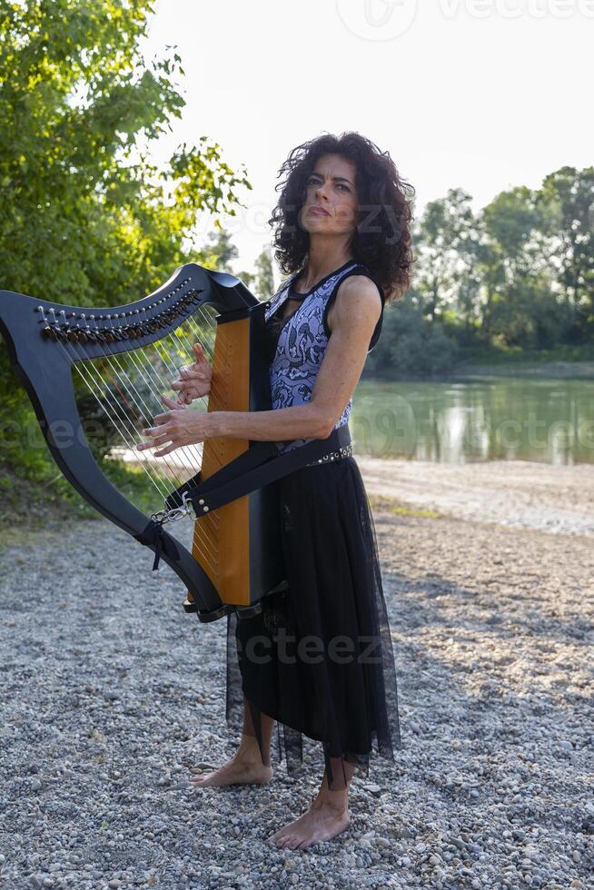 beautiful curly haired woman in fairytale clothes playing a small harp relaxing on a beach along the river at sunset photo