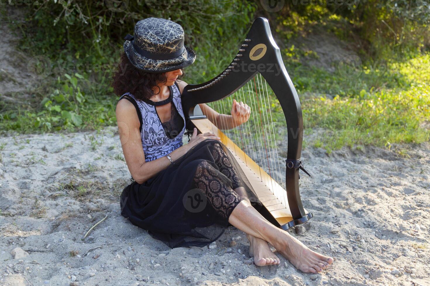 beautiful curly haired woman in fairytale clothes playing a small harp relaxing on a beach along the river at sunset photo