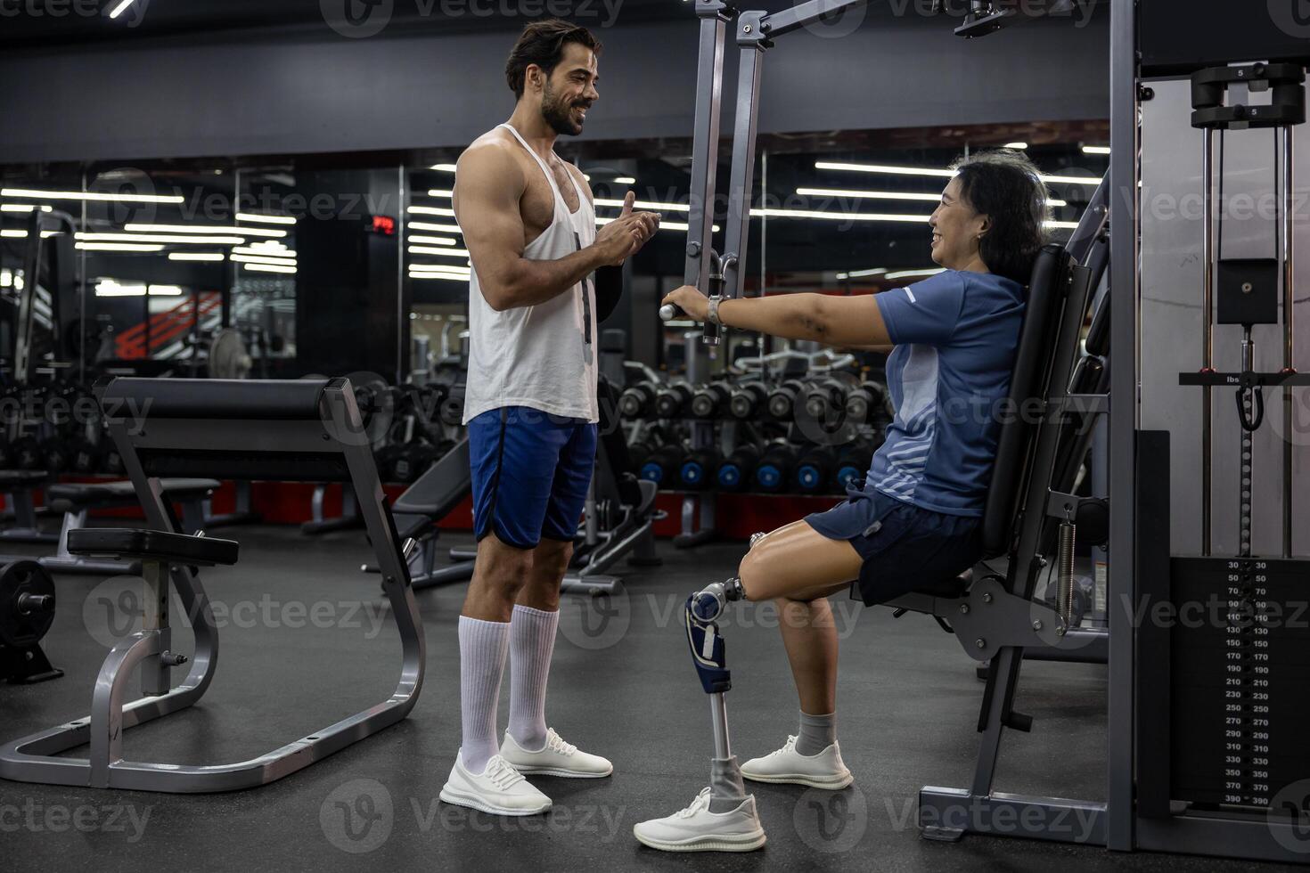 Asian woman with prosthetic leg from amputation from doing weight training workout exercise using machine at gym with professional trainer for handicap and disability people and recovery photo