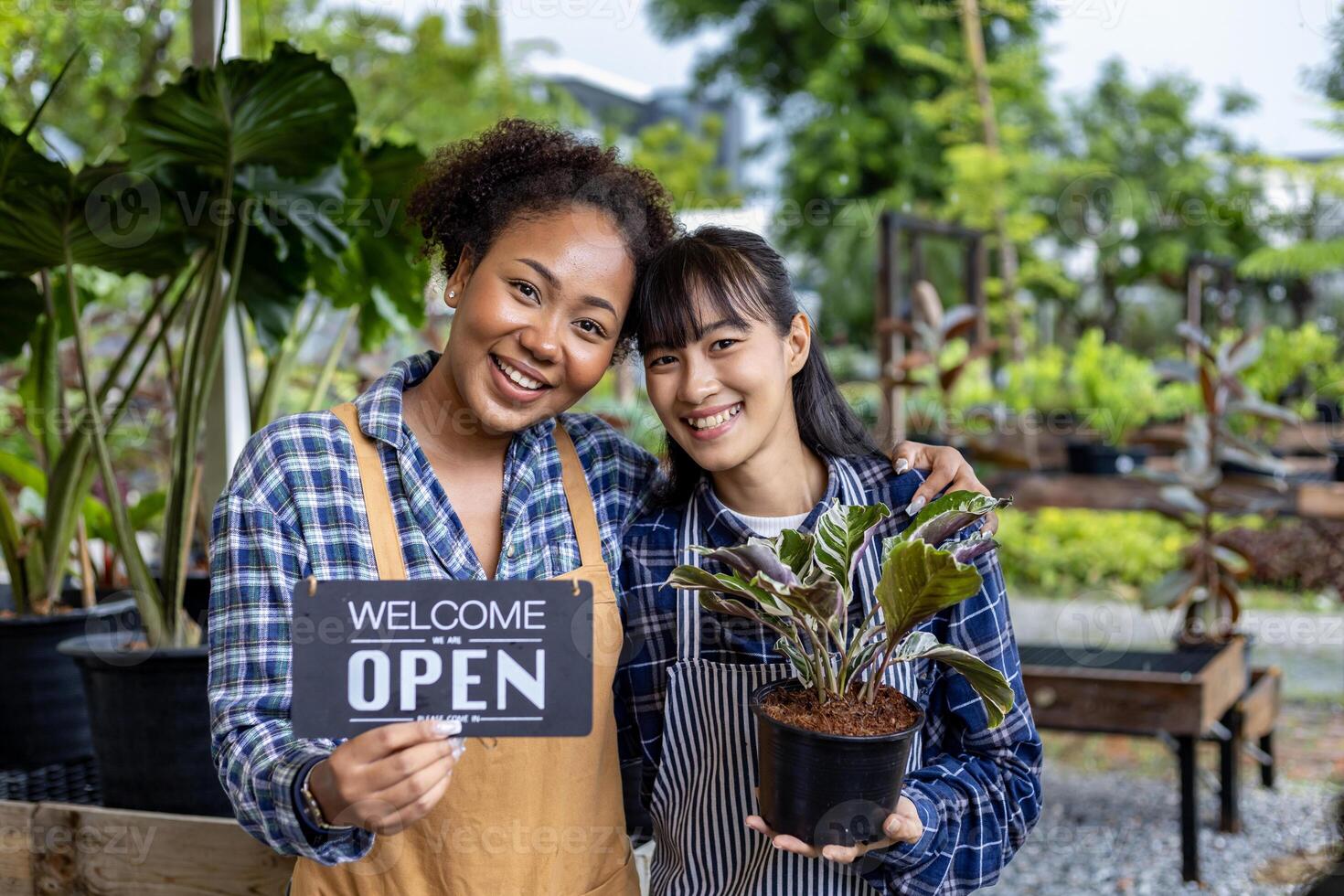 African gardener and owner with her asian partner are holding open sign in front of their ornamental houseplant at their own nursery garden center full of exotic and tropical plants photo