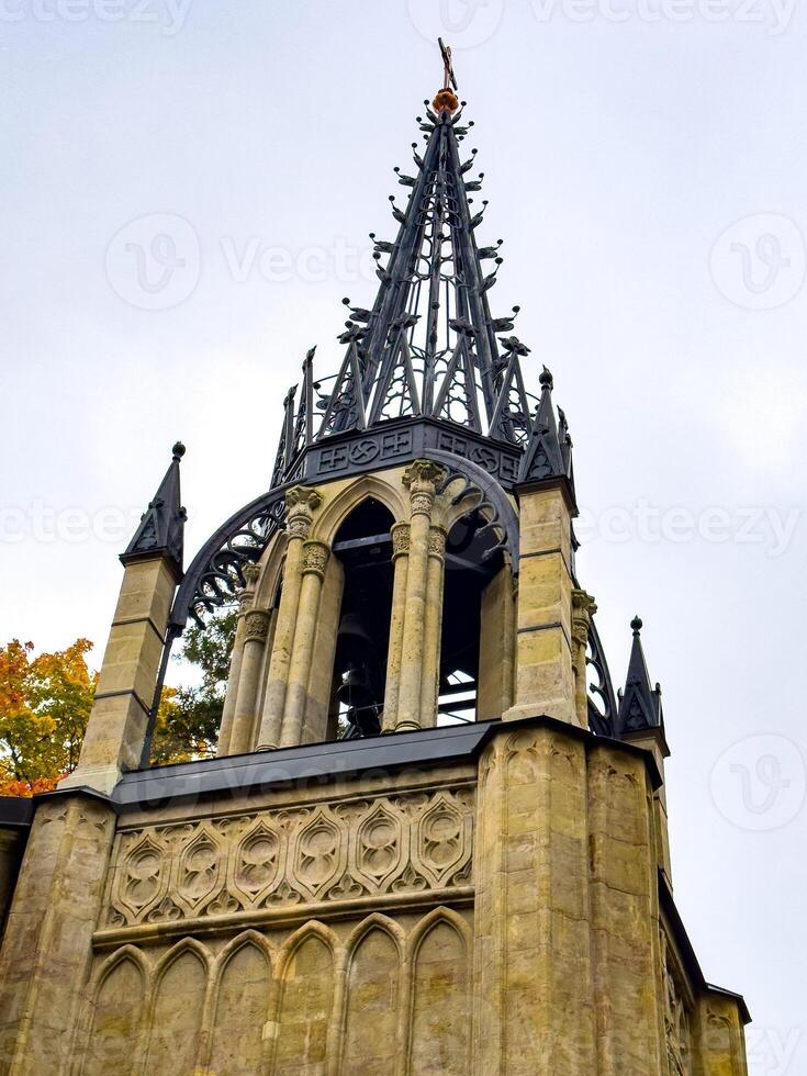 An imposing Gothic-style chapel with a tall, intricately detailed spire reaching into the overcast sky. The structure stands with its arched entrance and stone facade, embodying historical photo