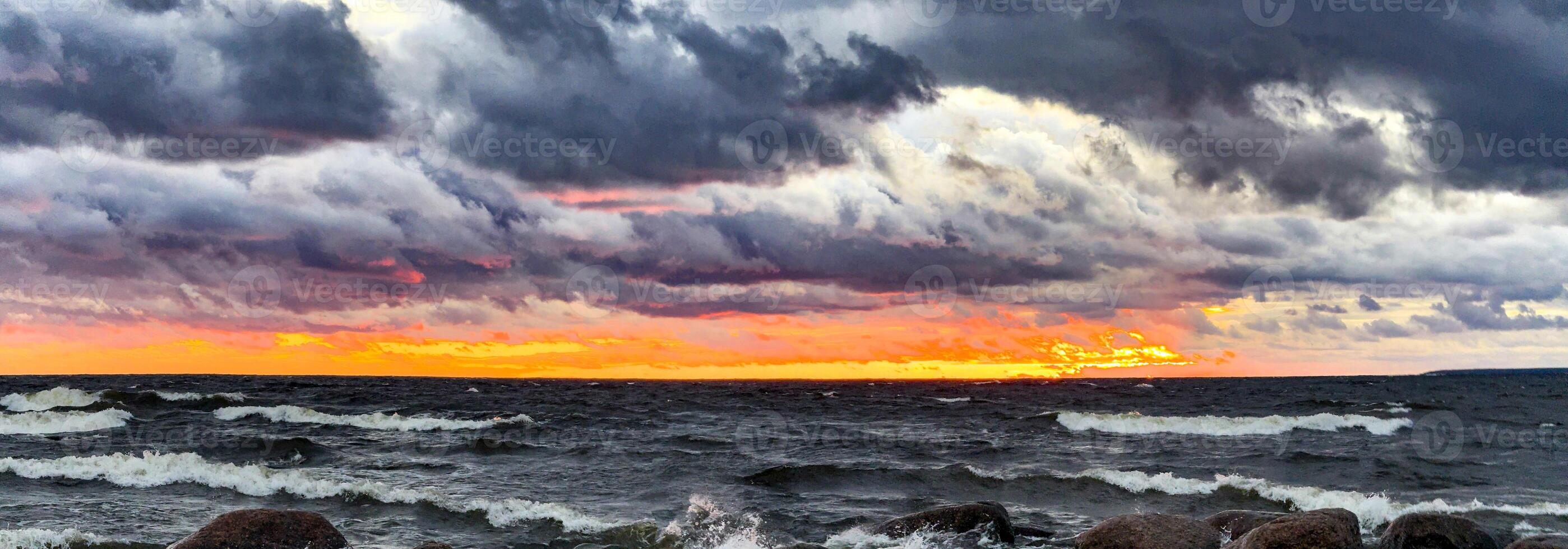 A breathtaking view of a rocky beach at sunset, with waves crashing against the stones under a dramatic sky. Dark clouds stretch across the horizon, while the setting sun casts a warm golden glow photo