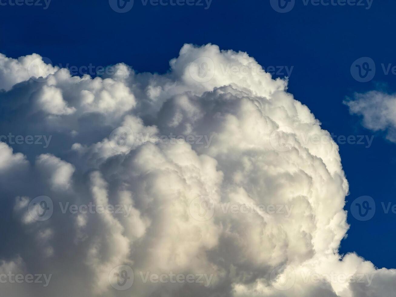 A breathtaking low-angle view capturing a towering cumulus cloud formation against a vivid blue sky. The sunlight enhances the cloud texture, highlighting the contrast between the bright white tops photo