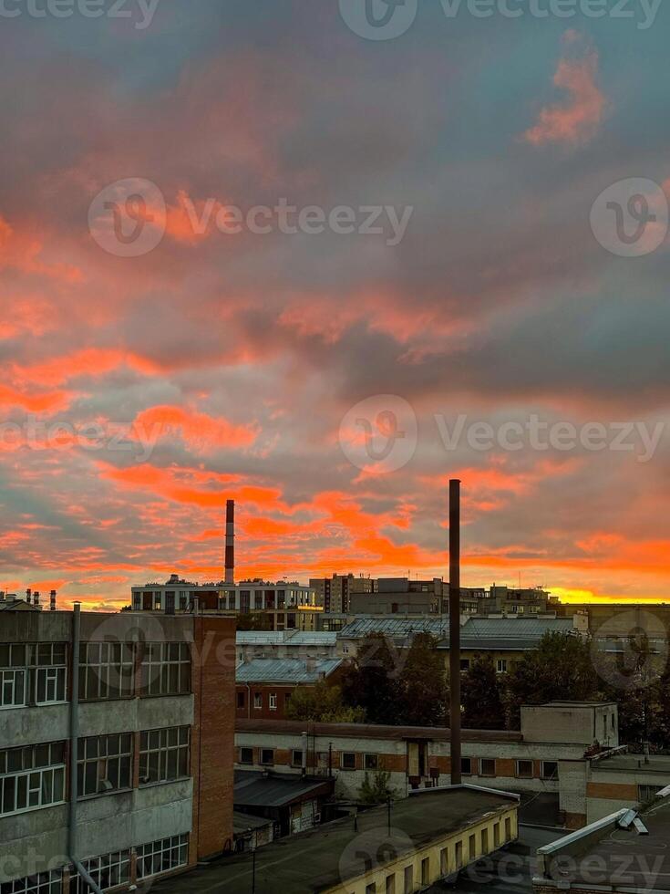 A captivating view of an industrial cityscape during a fiery sunset, where the sky is illuminated in intense shades of red and orange. The chimneys and rooftops create a sharp contrast against the photo