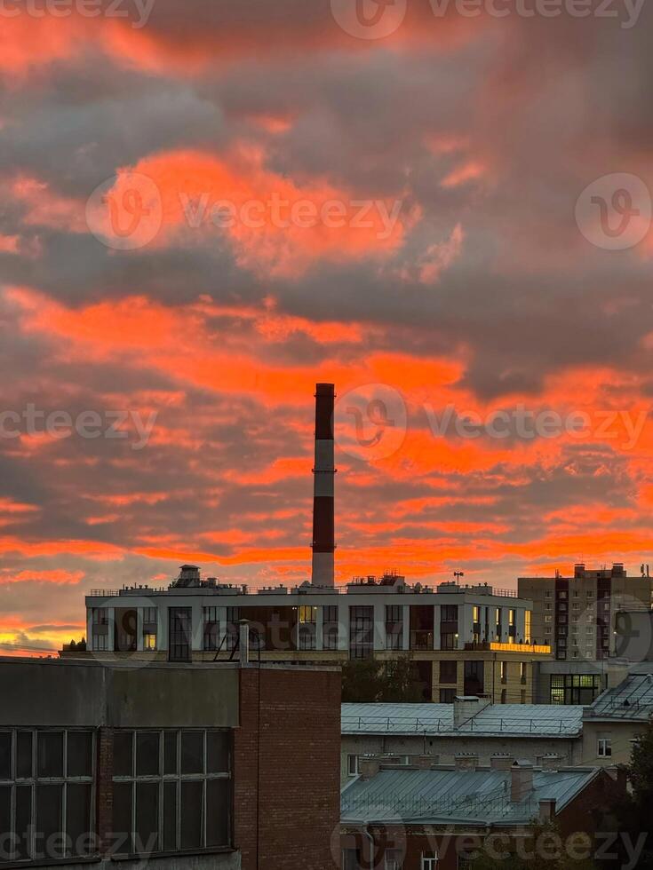 A captivating view of an industrial cityscape during a fiery sunset, where the sky is illuminated in intense shades of red and orange. The chimneys and rooftops create a sharp contrast against the photo