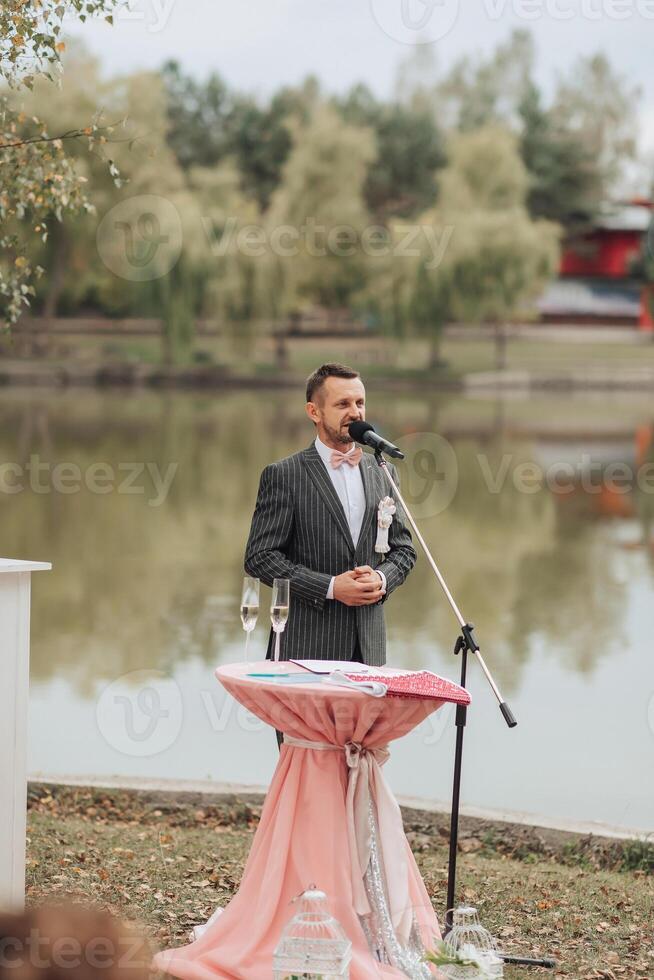 A man stands at a podium in front of a lake, singing into a microphone photo