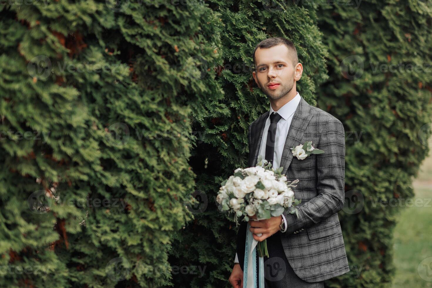 A man in a suit and tie stands in front of a bush holding a bouquet of flowers photo
