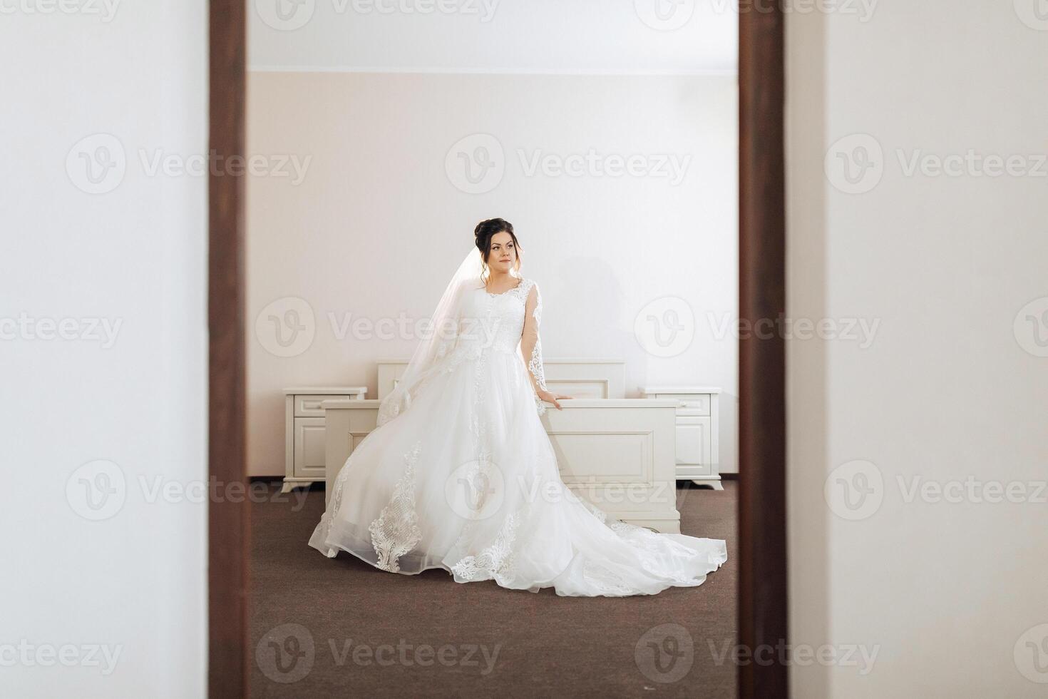 A bride is standing in front of a bed, wearing a white dress and a veil photo