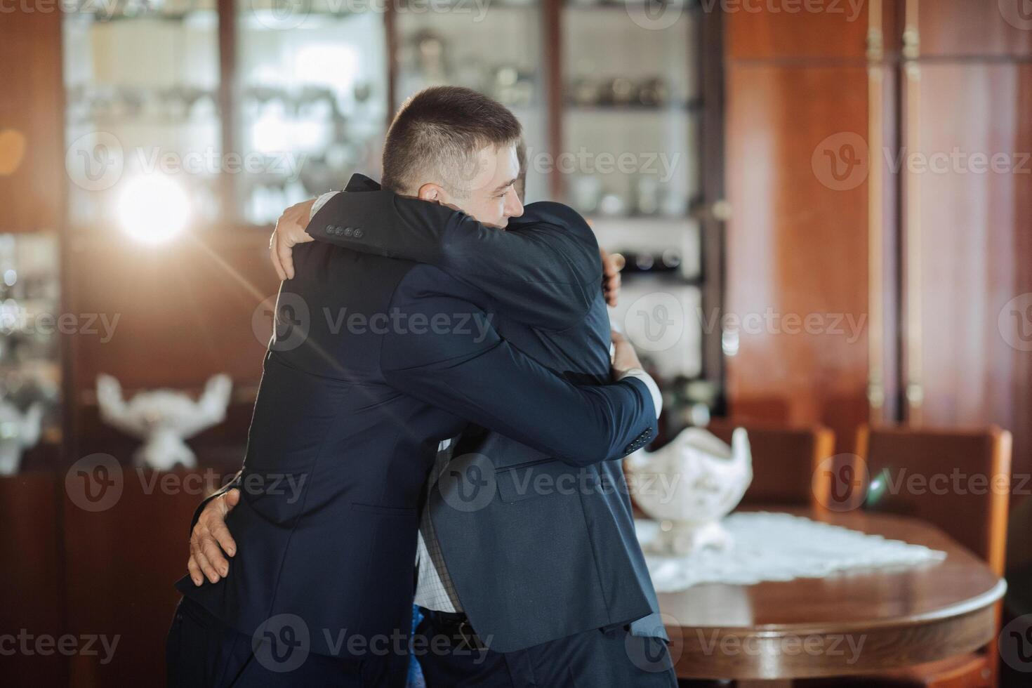 Two men hug each other in a room with a table in the background photo