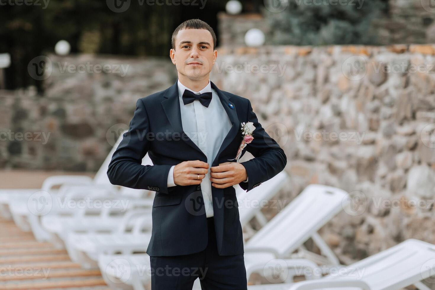 A man in a suit is standing in front of a stone wall, adjusting his tie photo