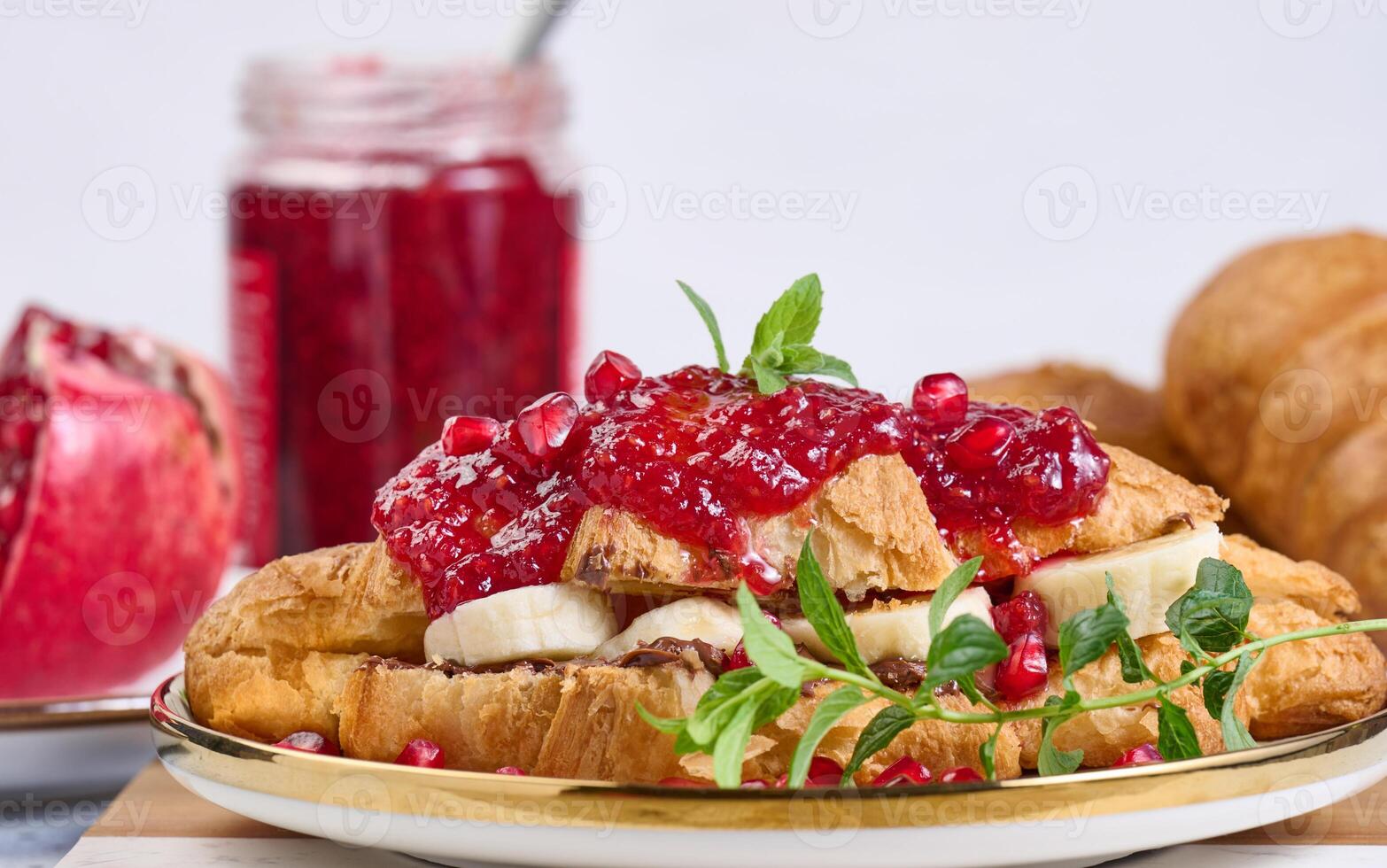Croissant with banana pieces, chocolate paste and pomegranate seeds in a round white plate photo