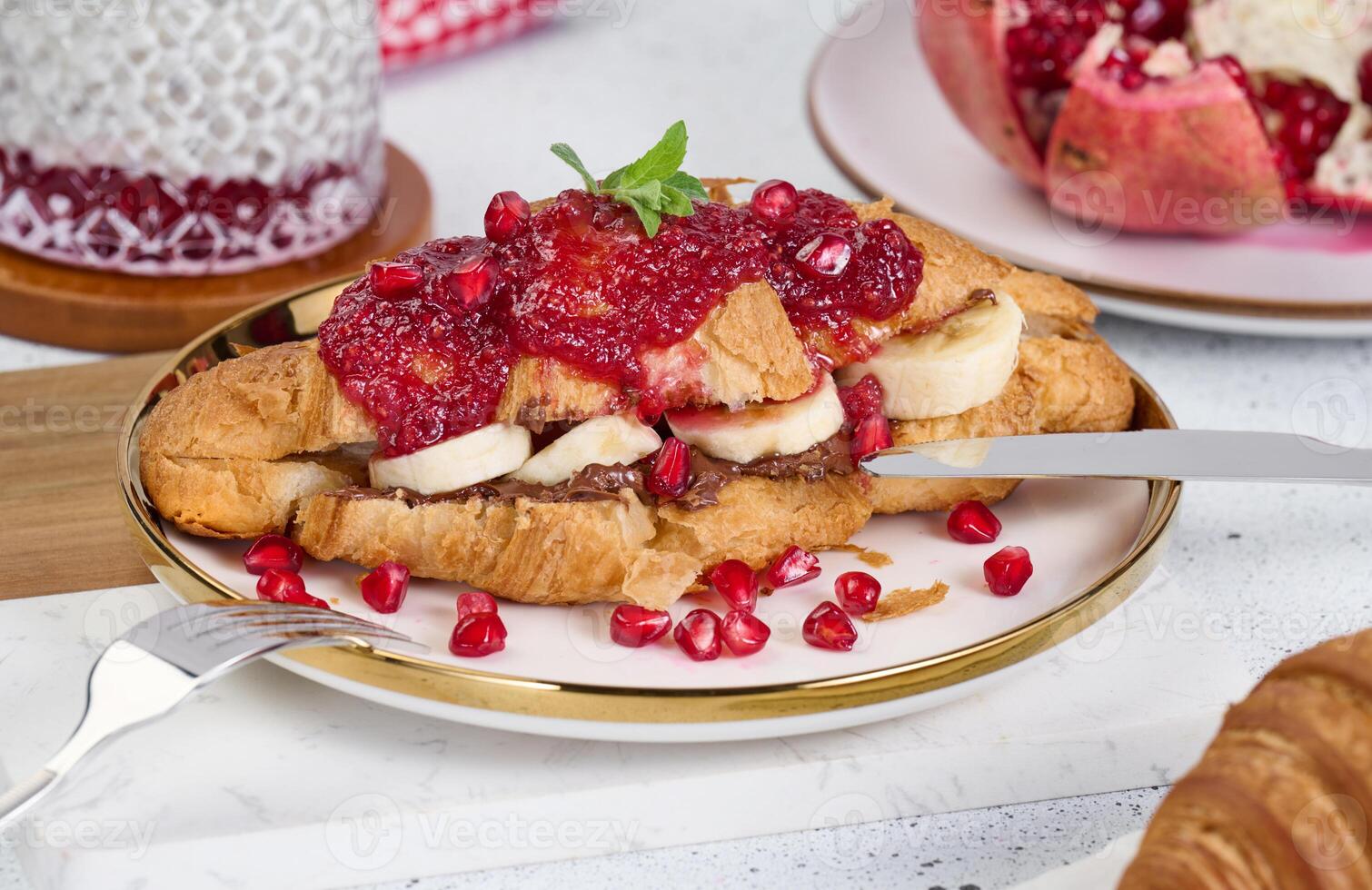 Croissant with banana pieces, chocolate paste and pomegranate seeds in a round white plate photo