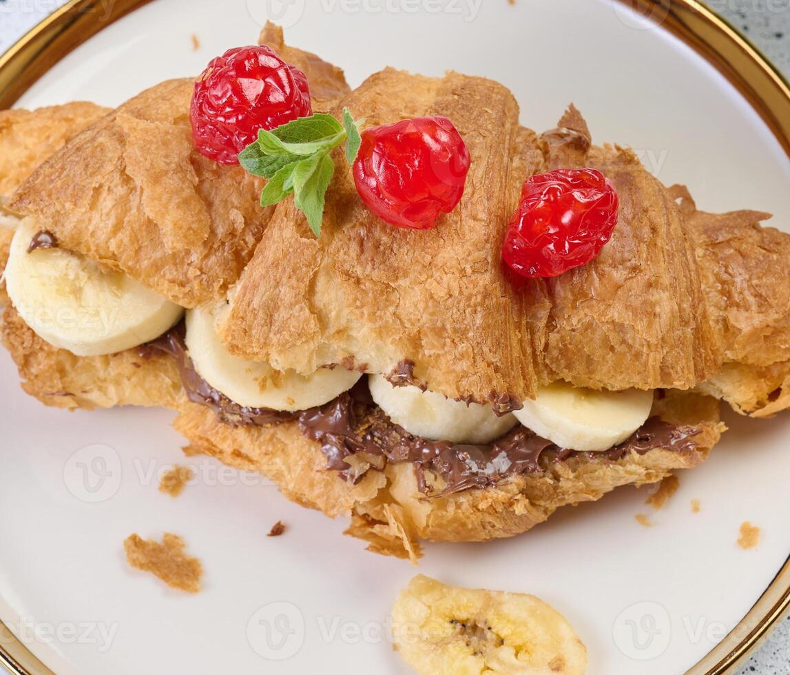 Croissant with chocolate spread and banana pieces in a white round plate on the table, breakfast photo