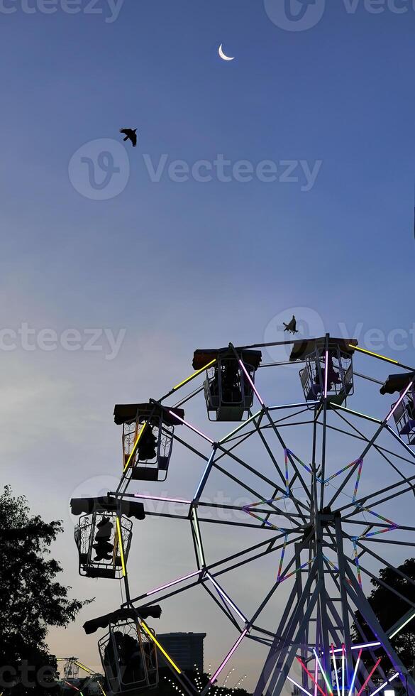 Low angle view of Ferris or Wonder wheel in silhouette shadow against twilight sky at Thai Red Cross Fair night market, amusement ride zone, carnival season photo