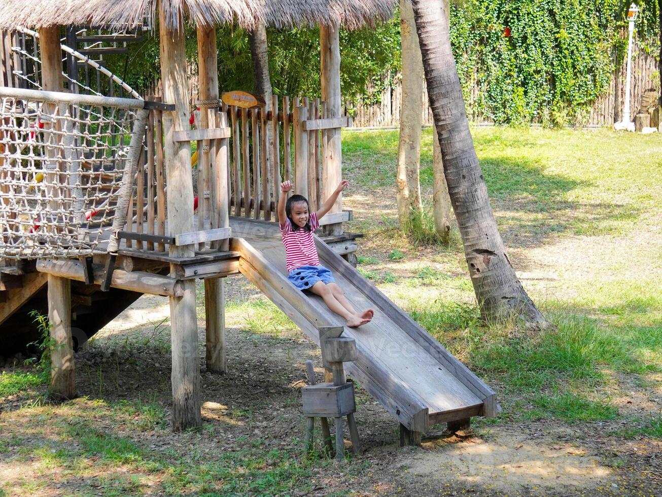 Children play outdoors on a wooden playground in a park. Happy girl playing on slide in playground in summer. photo